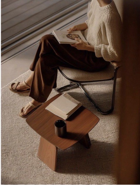 Reading in a quiet, warmly styled corner — knit sweater, wooden side table, and textured rug